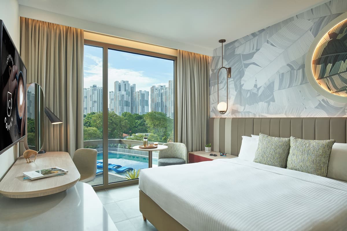 A hotel room with a large bed, a desk, a chair, a small table with a drink, and a view of a cityscape and pool.