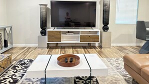 Een moderne woonkamer met een flatscreen televisie, houten en witte opbergmeubels, een salontafel met afstandsbedieningen en een kamerbreed tapijt met dessin.