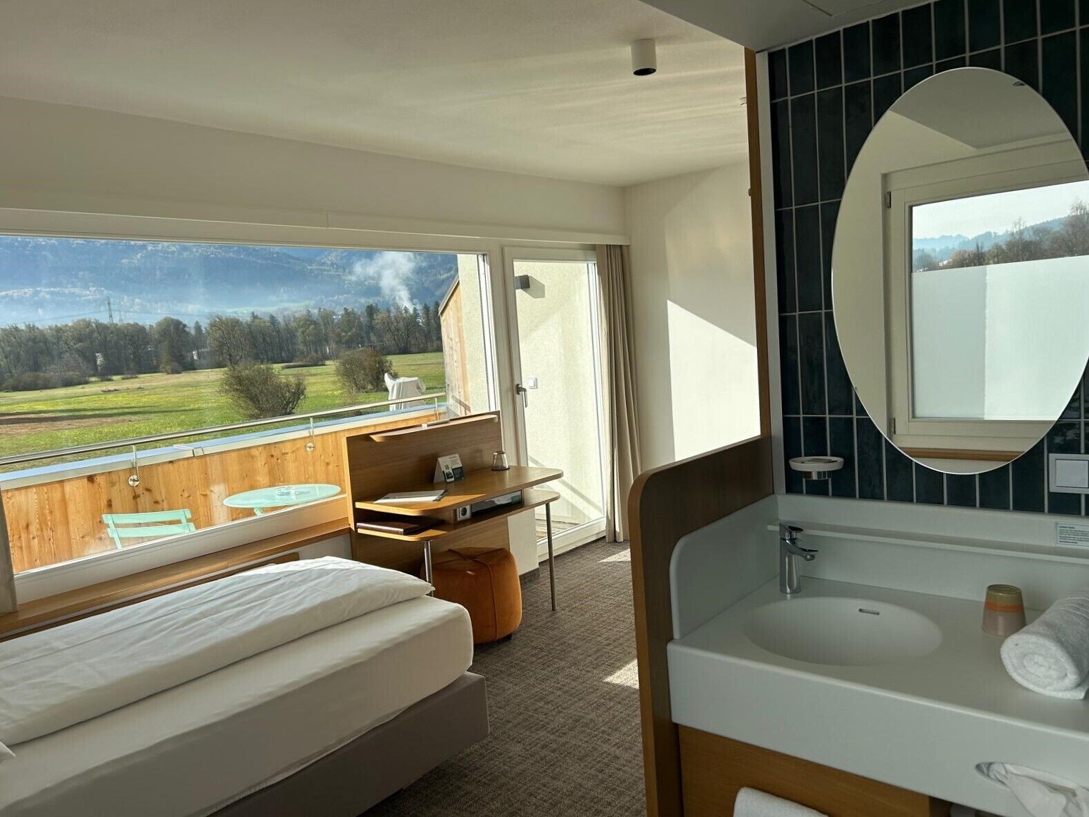 A hotel room with a bed, a bathroom with a large mirror, a sink, and a view of a green landscape through a large window.