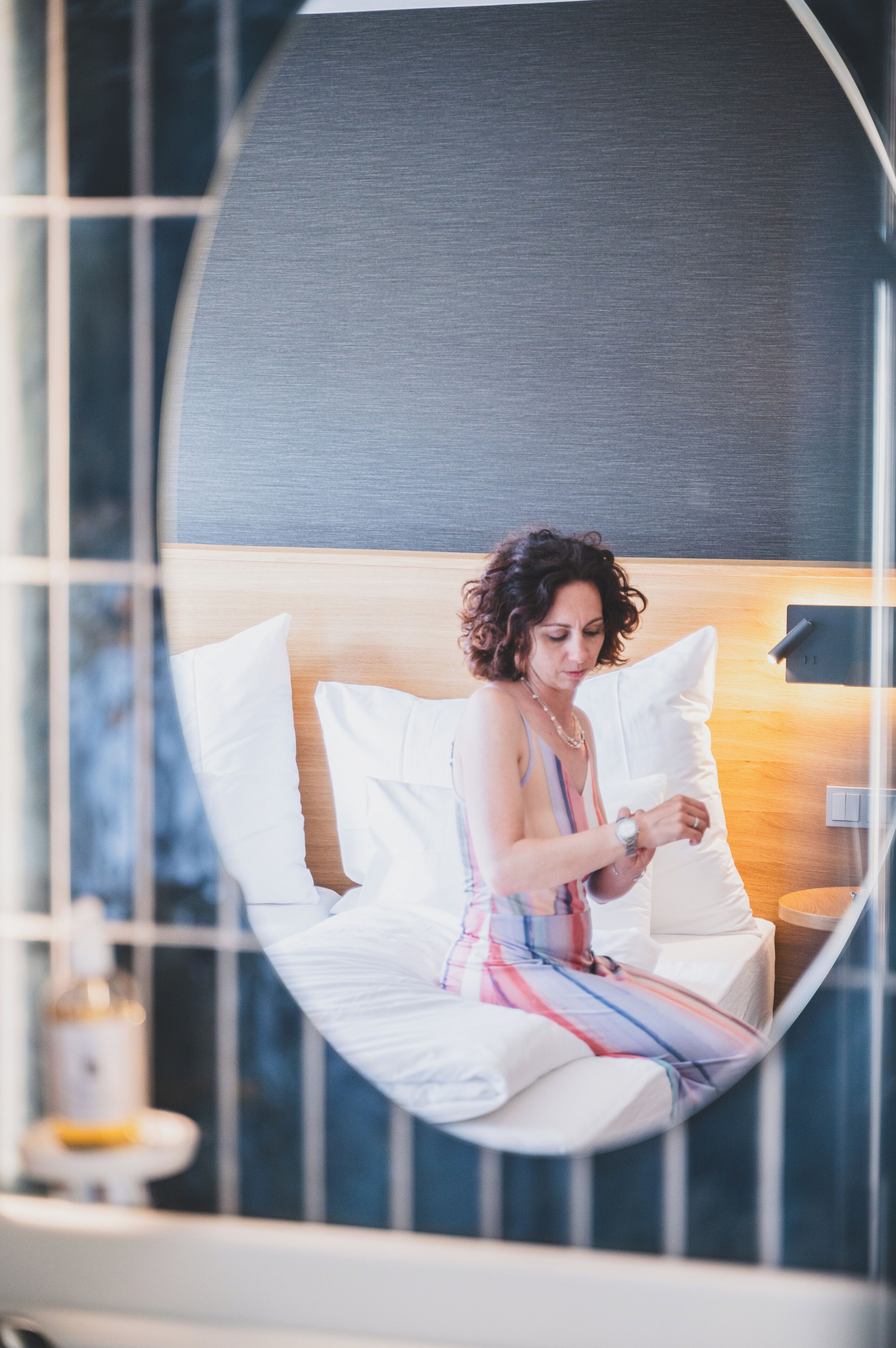 A woman sitting on a bed in a room with a large mirror, a wooden headboard, and a small table with a bottle.