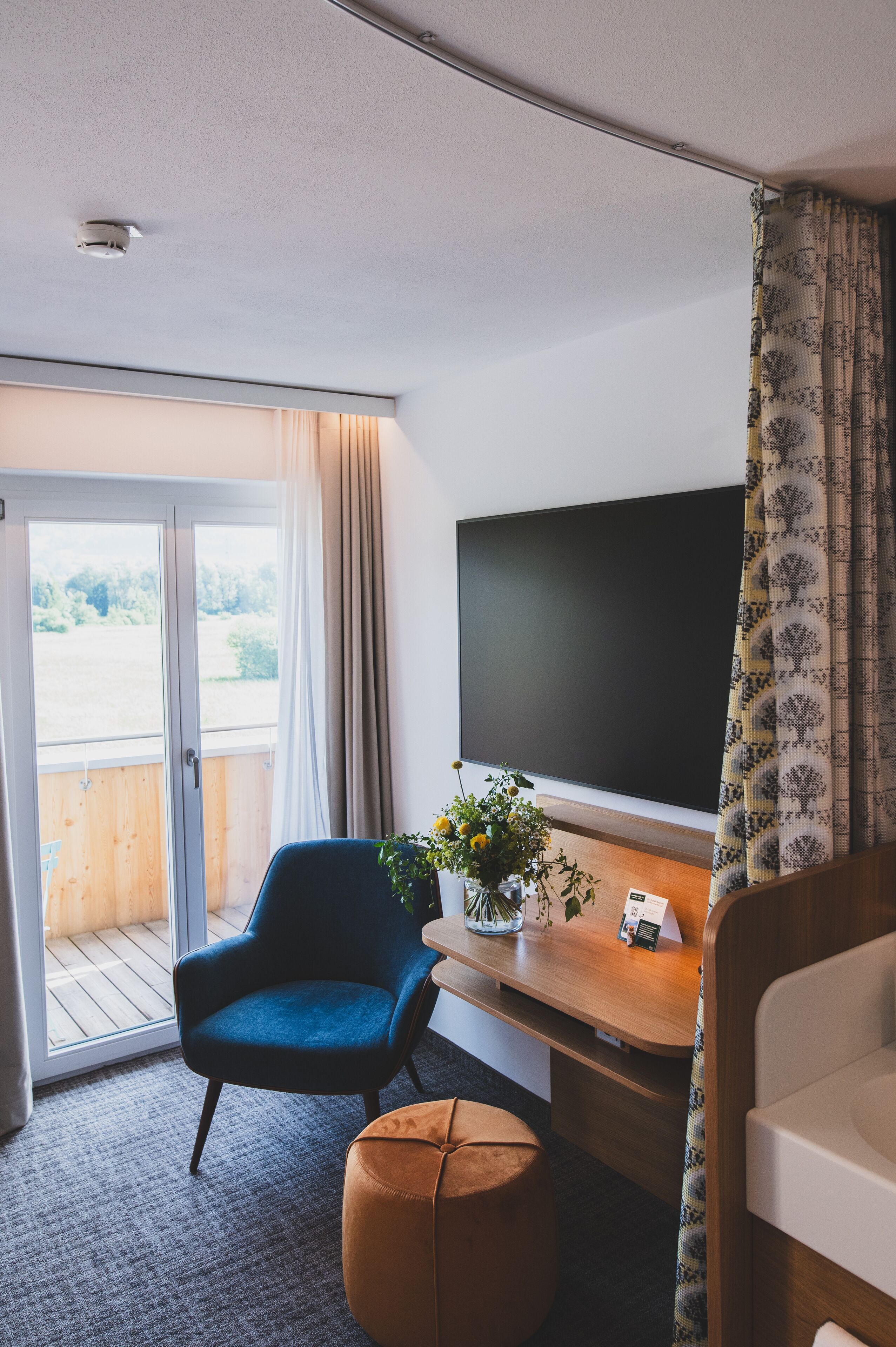 A modern room with a large window, a blue chair, a wooden desk, a television, and a small potted plant.