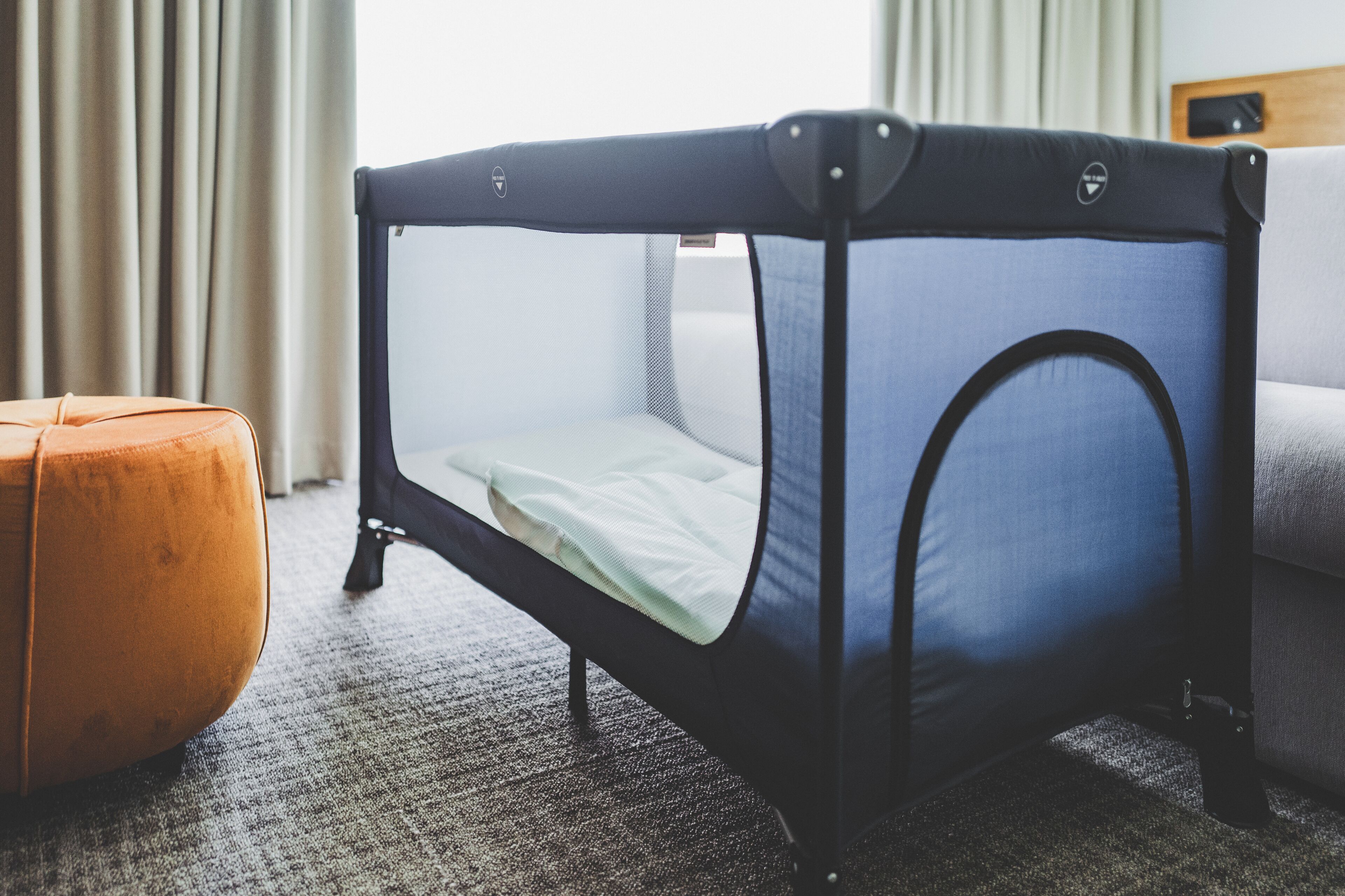 A baby crib with a mesh top and a mattress, positioned next to a grey sofa and an orange ottoman in a room with a carpeted floor.