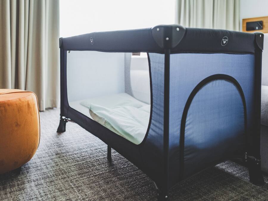 A baby crib with a mesh top and a mattress, positioned next to a grey sofa and an orange ottoman in a room with a carpeted floor.