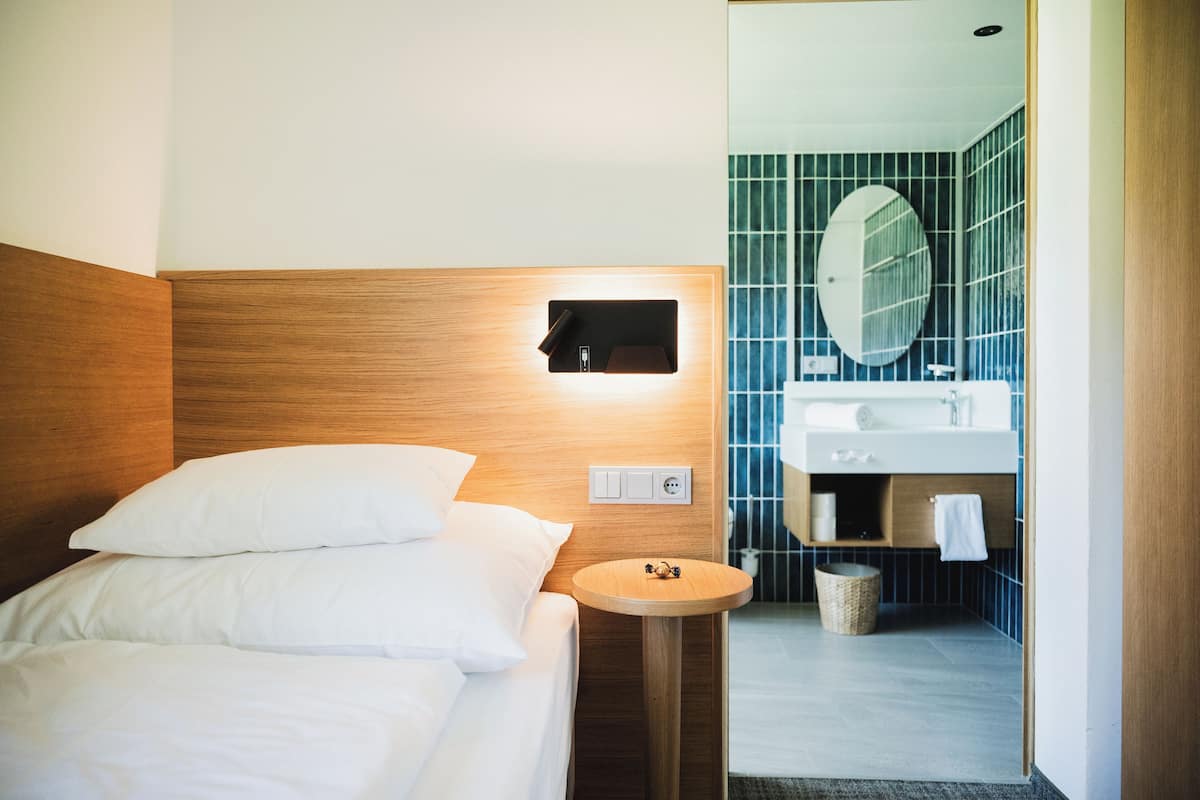 Habitación de hotel con cabecera de madera, una mesita de noche redonda de madera, cama con ropa blanca, luz empotrada en la pared, baño con espejo redondo, lavabo y un perchero para toallas.