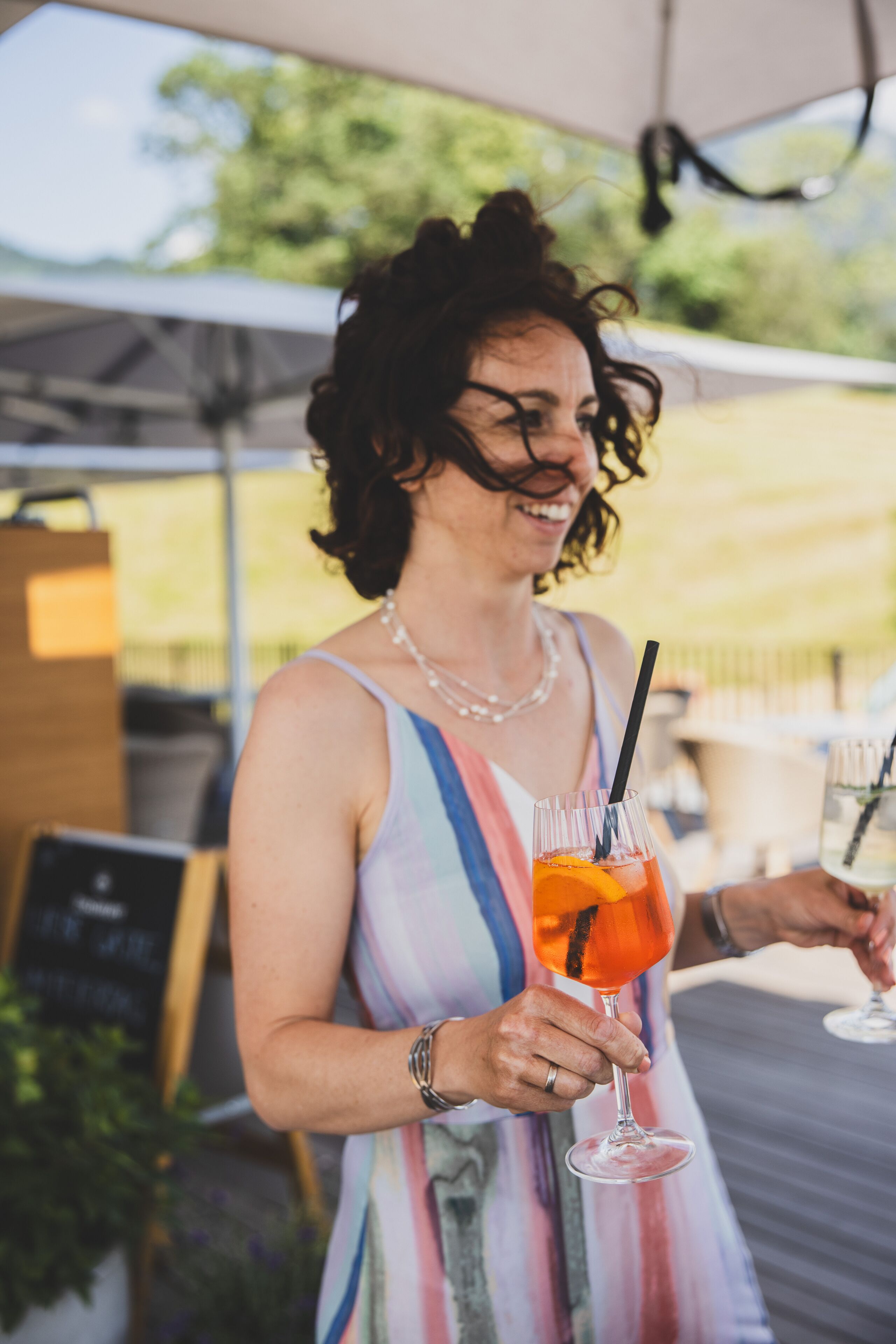 A person holding a glass with a straw, wearing a striped dress, and standing under a tent.