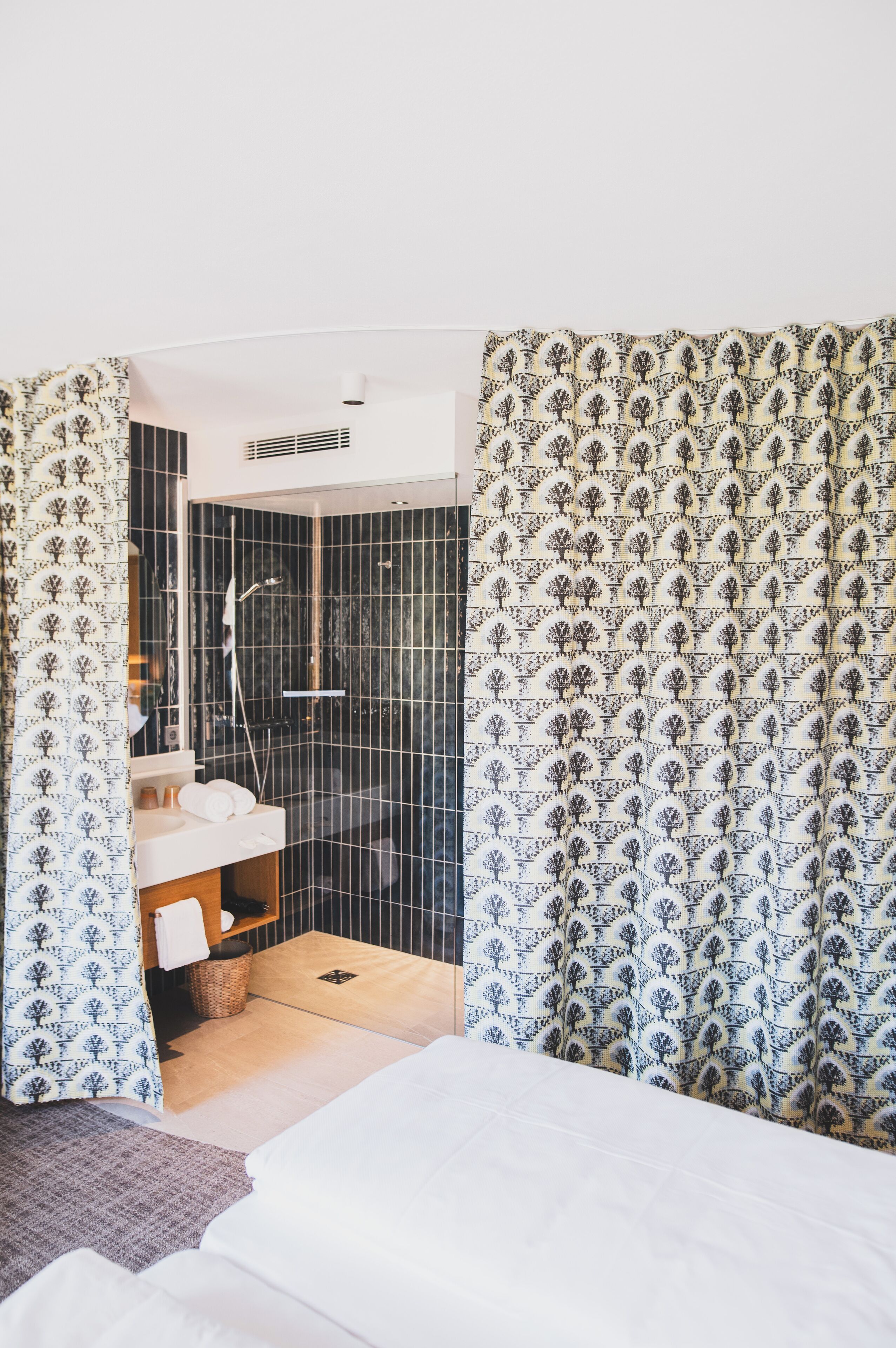 A modern hotel room with a shower area, a white bed, and patterned curtains.