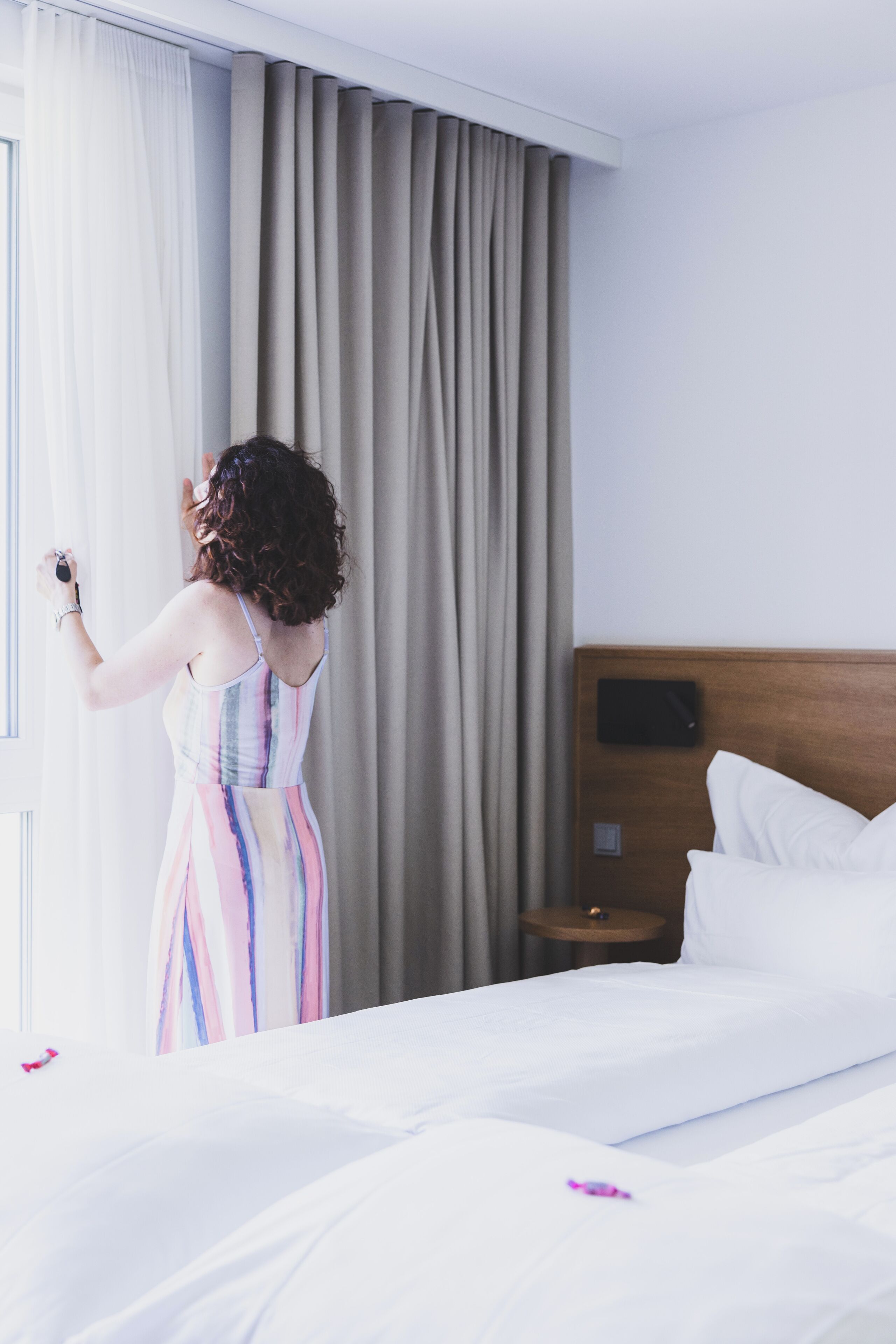 A hotel room with a bed, a nightstand, a window with curtains, and a view of the outdoors.
