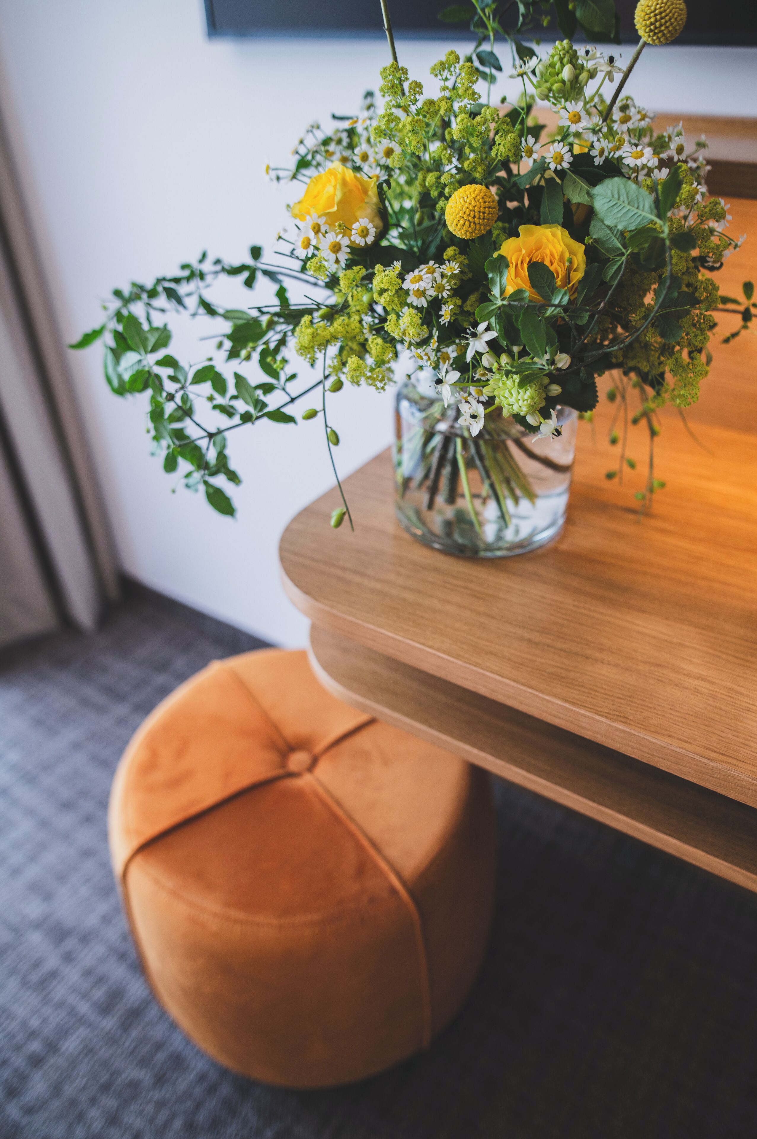 Una mesa de madera con un florero de vidrio transparente que contiene flores amarillas y follaje verde.