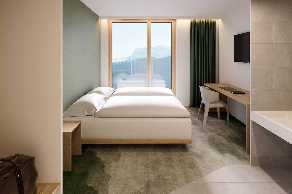 Habitación de hotel moderna con cama, escritorio y vistas a montañas a través de amplios ventanales.