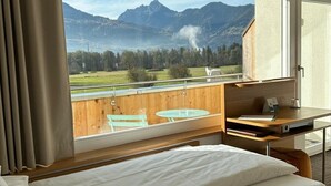 A hotel room with a large window, a bed, a desk, and a view of mountains.