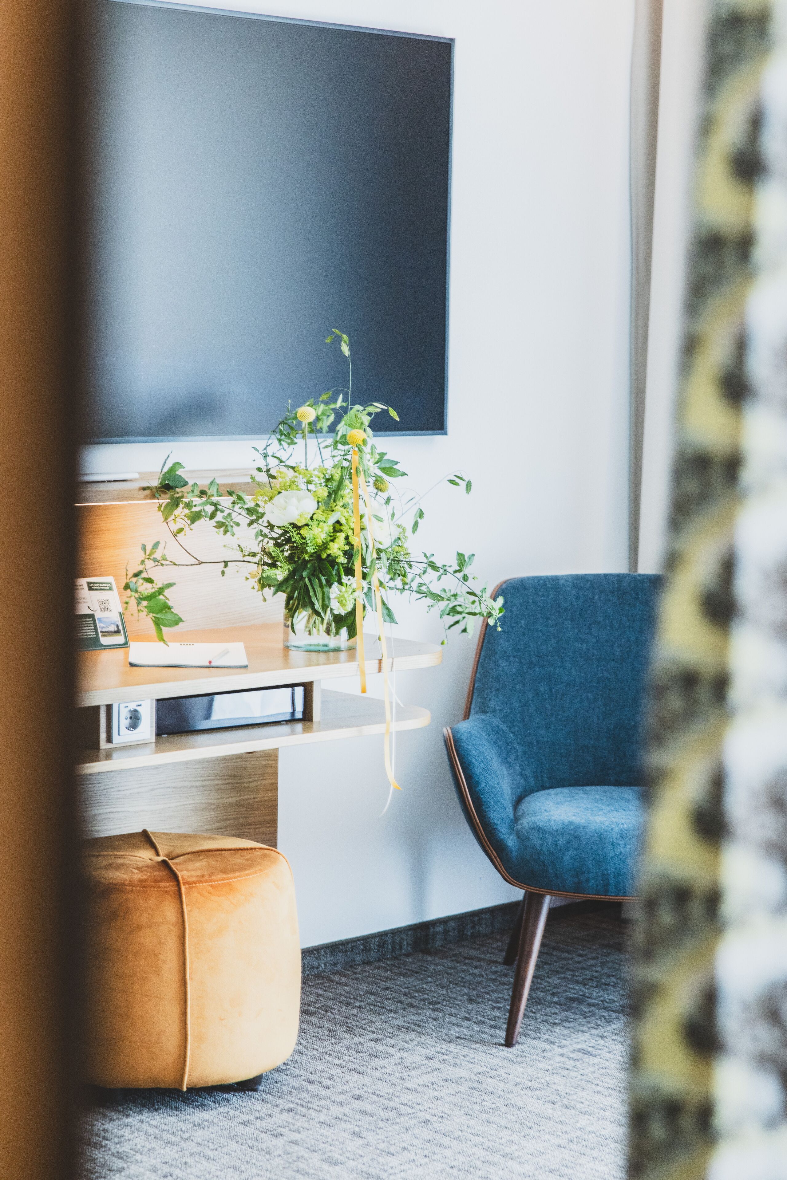 A modern room with a desk, a blue chair, a wooden ottoman, and a potted plant.