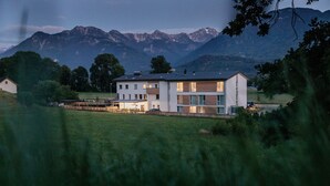 A modern two-story house with a flat roof, surrounded by a grassy field and mountains in the background.