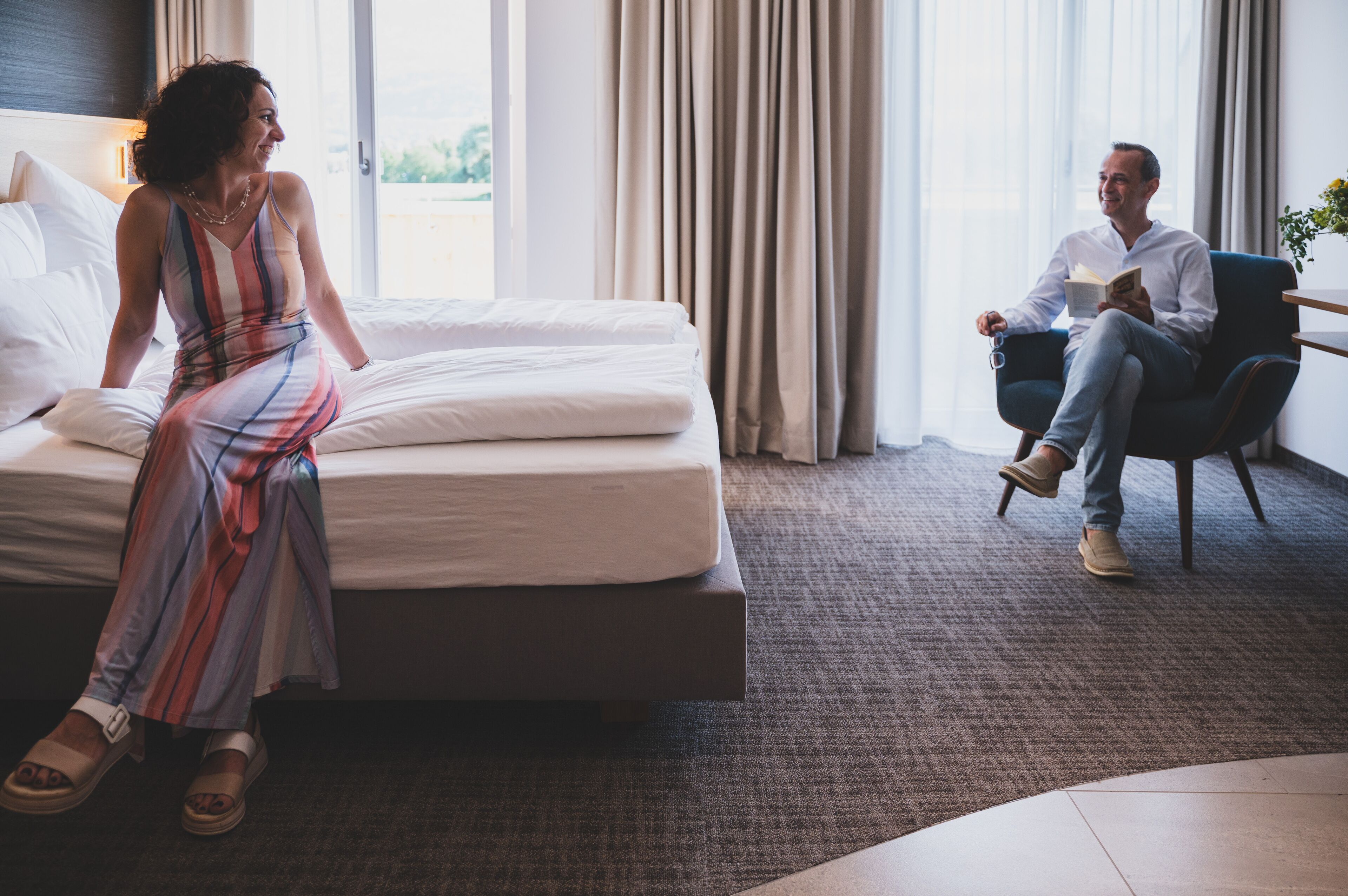 A hotel room with a bed, a sitting area with a chair, and a large window with curtains.