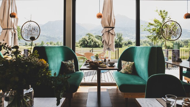 A dining area with green upholstered chairs, a table with a bowl of fruit, and a view of mountains through large windows.
