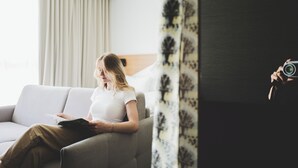 A woman sitting on a grey sofa in a room with a window and curtains.