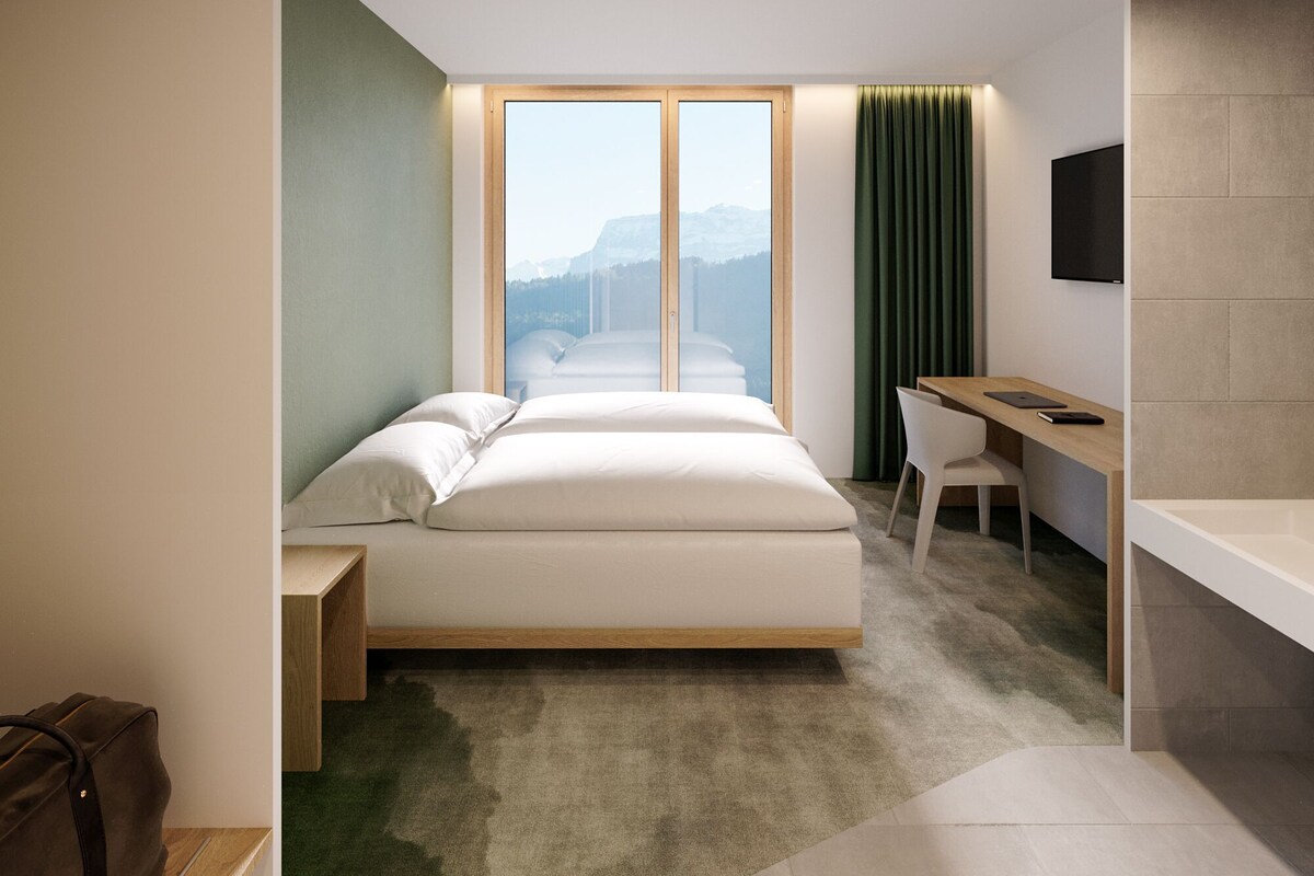 Une chambre d’hôtel moderne, équipée d’un lit, d’un bureau et offrant une vue sur les montagnes grâce à de grandes fenêtres.
