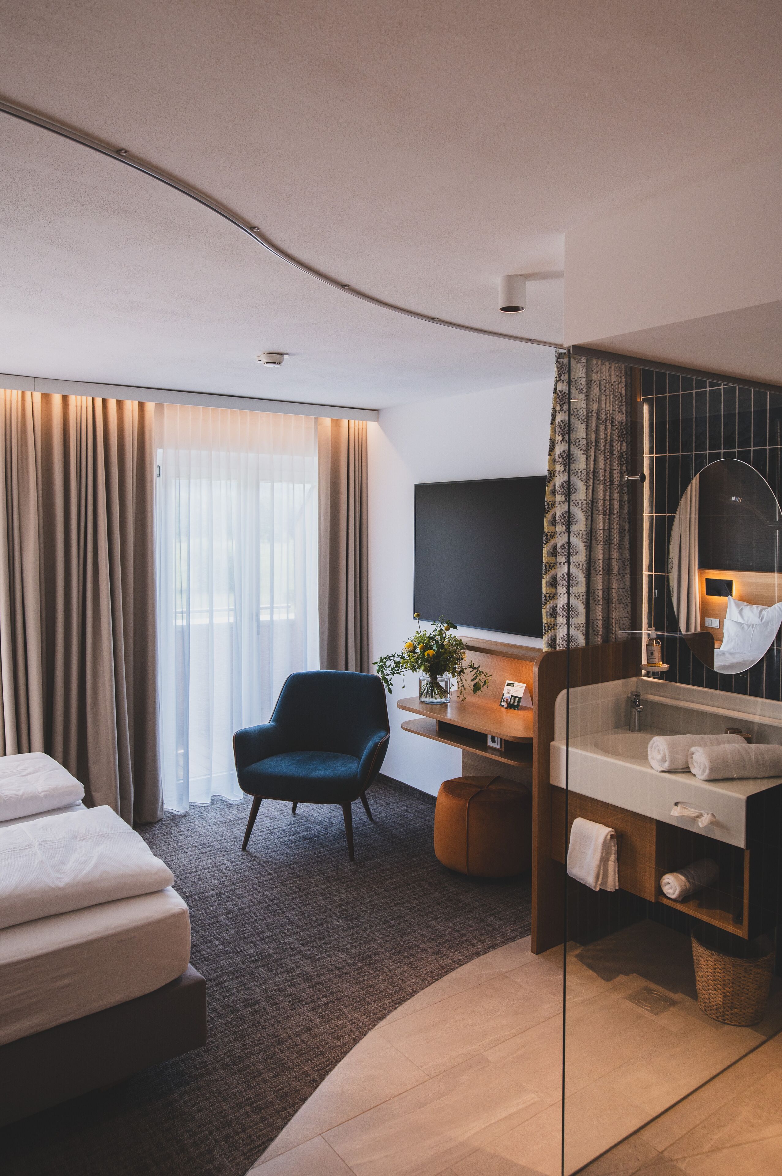 A modern hotel room with a bed, a desk with a computer, a chair, a bathroom with a mirror, and a TV.