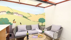 Un patio acogedor con mobiliario de mimbre, una mesa con una botella y copas, y un mural de un paisaje.