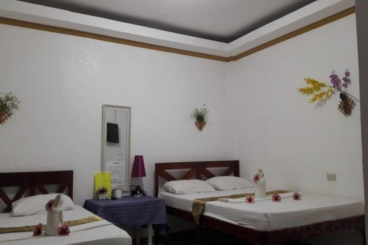 A room with two beds, a small table with a lamp, and a wall-mounted shelf with decorative items.