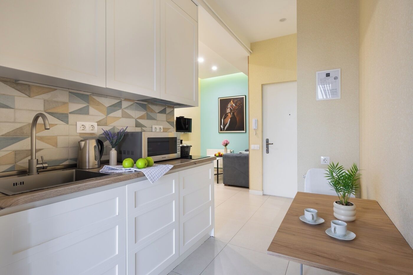 A modern kitchen with white cabinets, a stainless steel sink, a microwave, and a coffee maker. There is a small dining area with a wooden table and two white cups.
