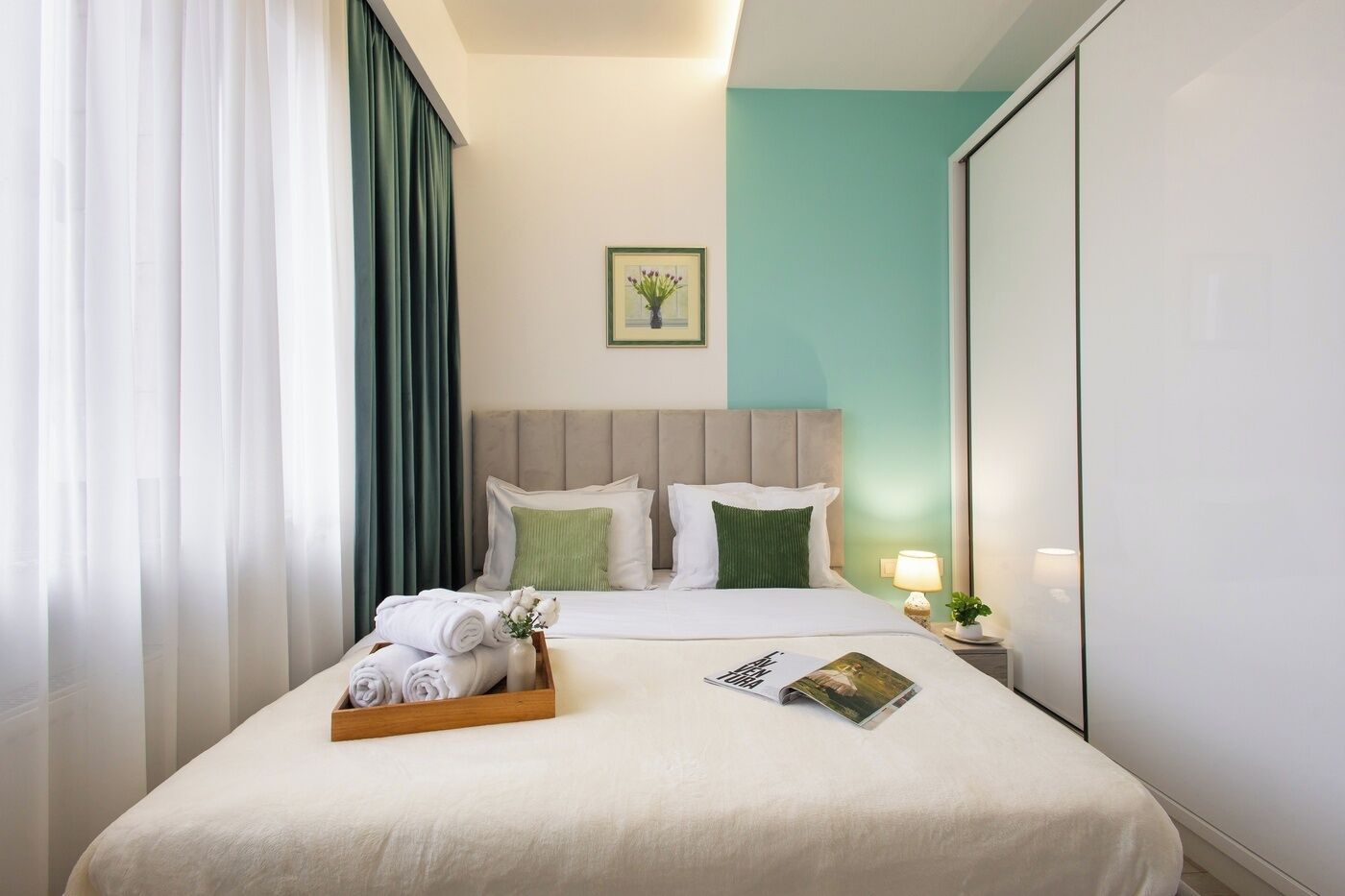 A hotel room with a bed, a nightstand with a lamp, a tray with towels, and a framed picture on the wall.