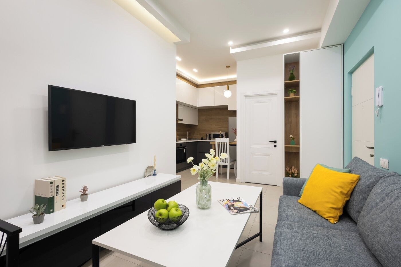 A modern living room with a flat-screen TV, a coffee table with a bowl of apples, a grey sofa with a yellow pillow, and a kitchen in the background.