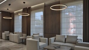 A modern lobby with sofas and armchairs arranged around a central coffee table, large windows with curtains, and pendant lighting. - Central Park Resident (Bishkek)