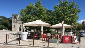 An outdoor café with red chairs and white umbrellas, a stone building in the background, and a paved area. - La dame de coeur (Soulac-sur-Mer)