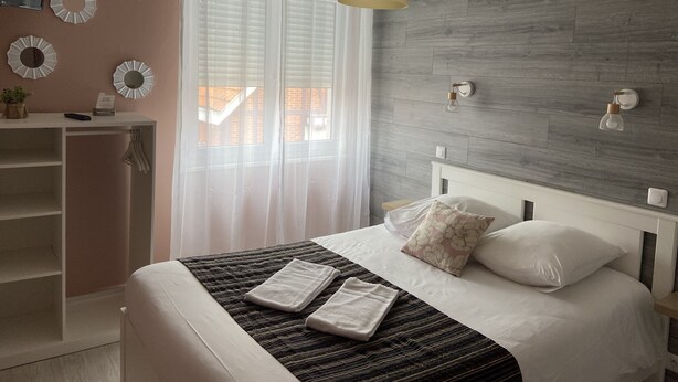 A bedroom with a bed, a bedside table, a mirror, and a window with curtains. - La dame de coeur (Soulac-sur-Mer)