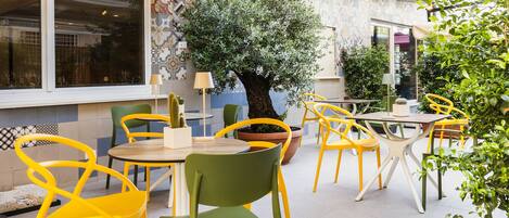 An outdoor seating area with yellow and green chairs, a large potted tree, and a patterned wall.