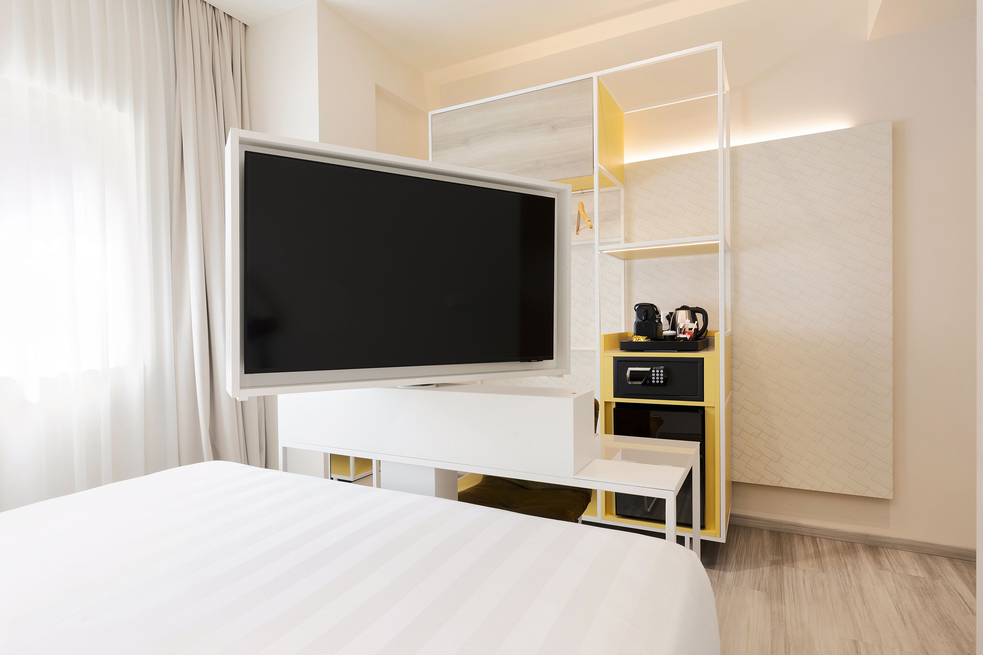 A modern hotel room with a large flat-screen TV on a white entertainment unit, a bed with a white headboard, and a built-in shelf with a coffee maker and other items.