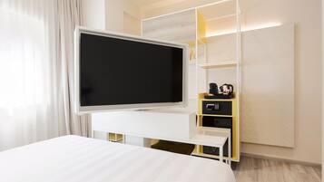 A modern hotel room with a large flat-screen TV on a white entertainment unit, a bed with a white headboard, and a built-in shelf with a coffee maker and other items.