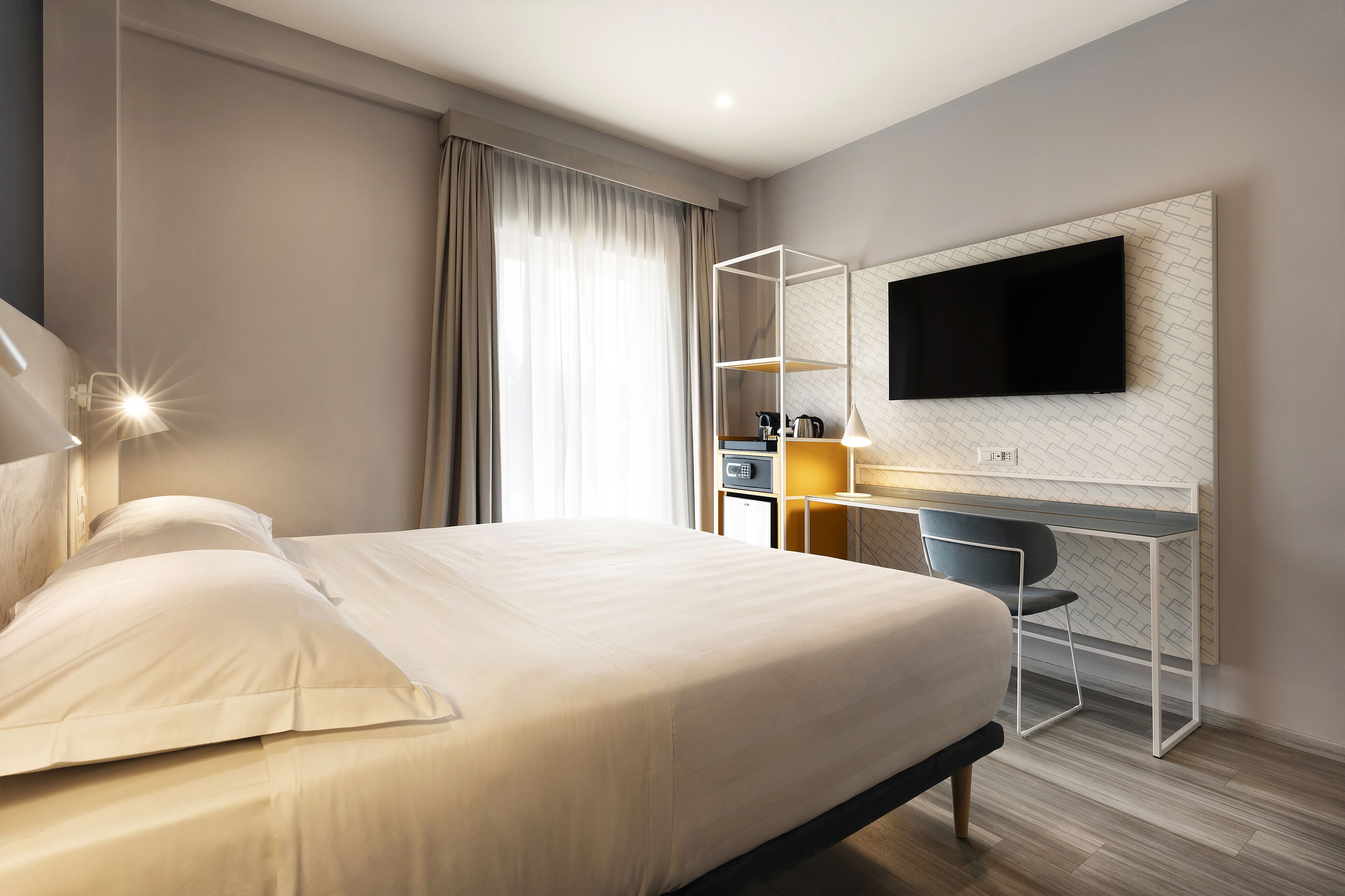 Une chambre d’hôtel moderne dotée d’un grand lit, d’un bureau avec une chaise, d’un téléviseur à écran plat fixé au mur et d’une petite table avec une lampe.