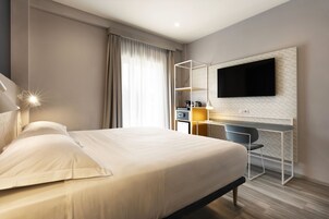 Una habitación de hotel moderna con una cama grande, un escritorio con una silla, un televisor de pantalla plana montado en la pared y una mesita de noche con una lámpara.