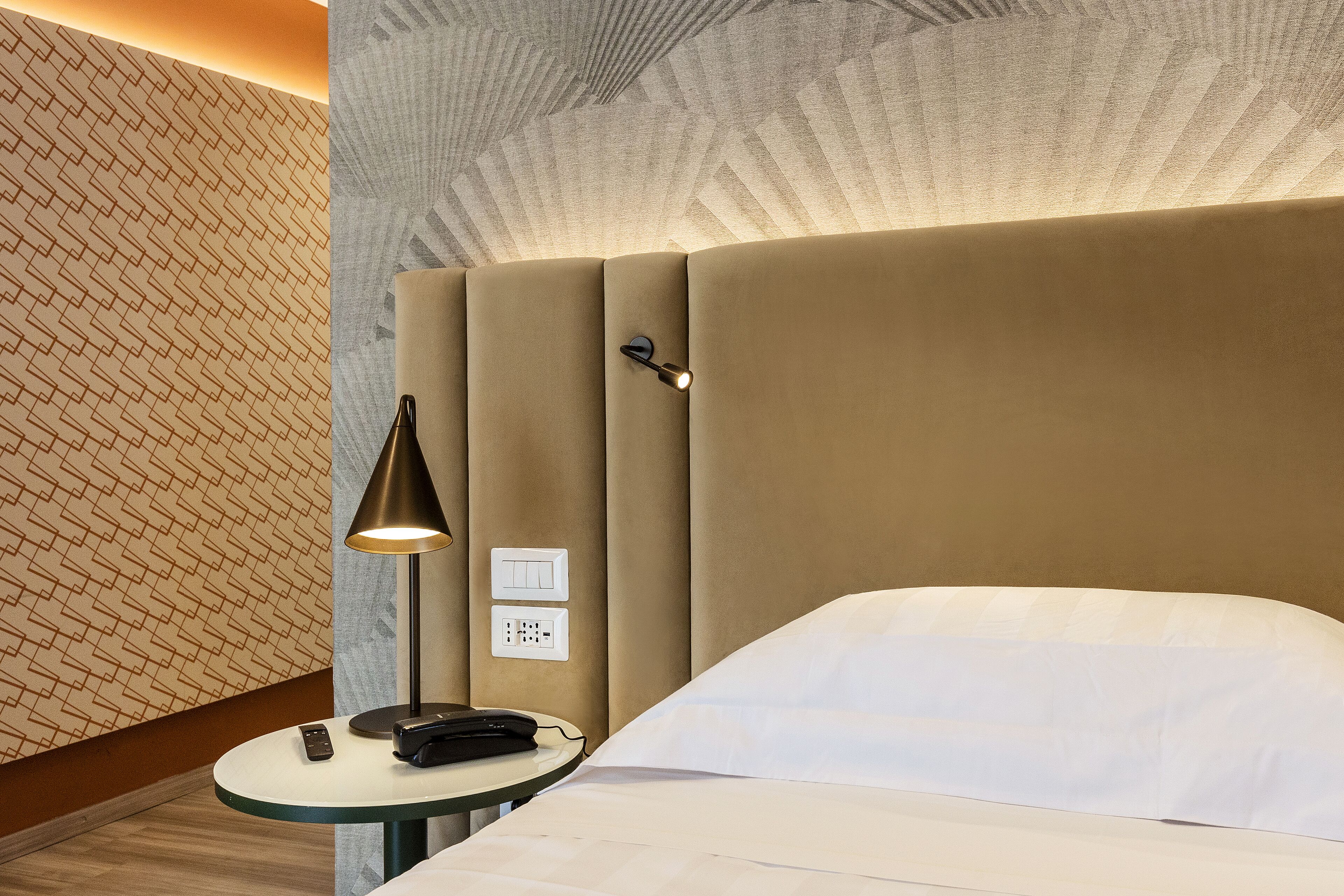 Une chambre d’hôtel avec un lit, une table de chevet avec une lampe, une télécommande et un mur orné d’un motif géométrique.