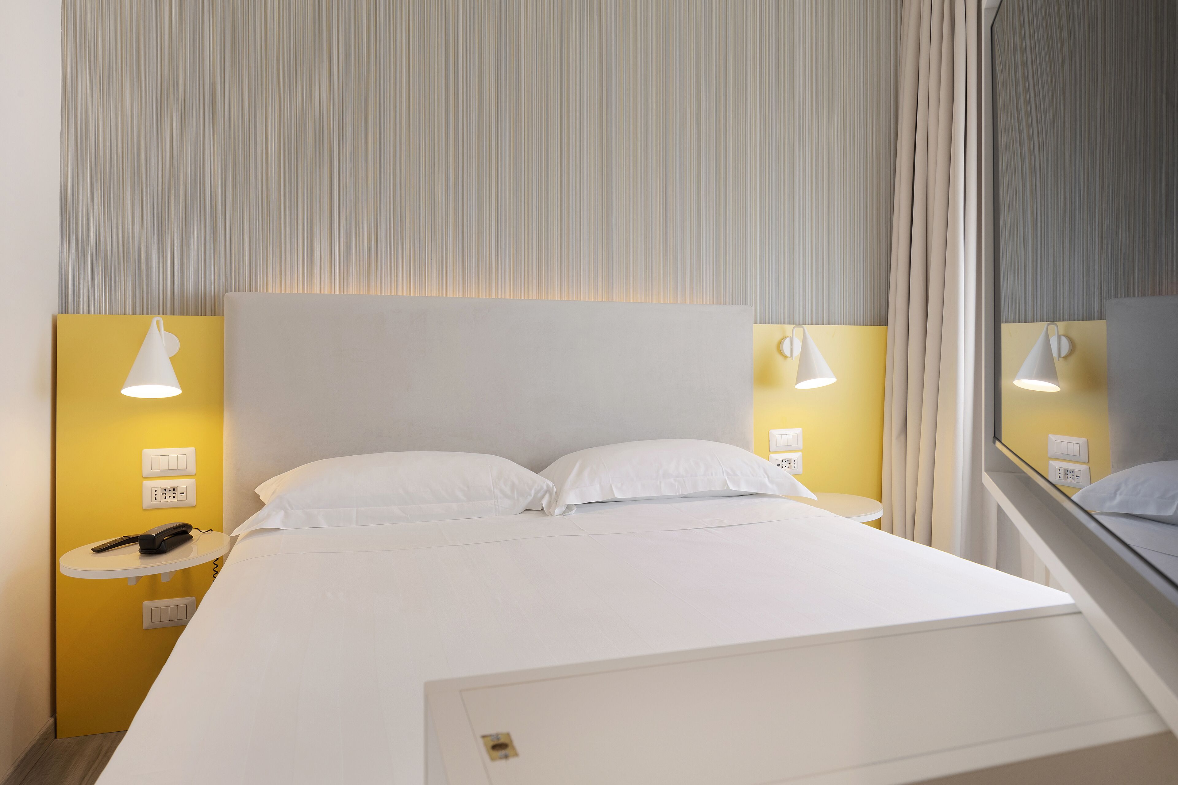 Una camera d'albergo con un letto grande, due abat-jour, un telefono sul comodino e una finestra con tende.