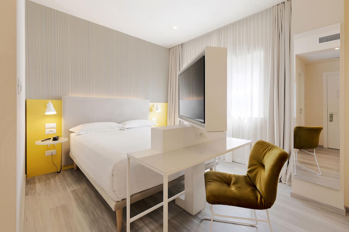 Habitación de hotel con cama, escritorio con computadora, silla, mesita de noche con lámpara y ventana con cortinas.