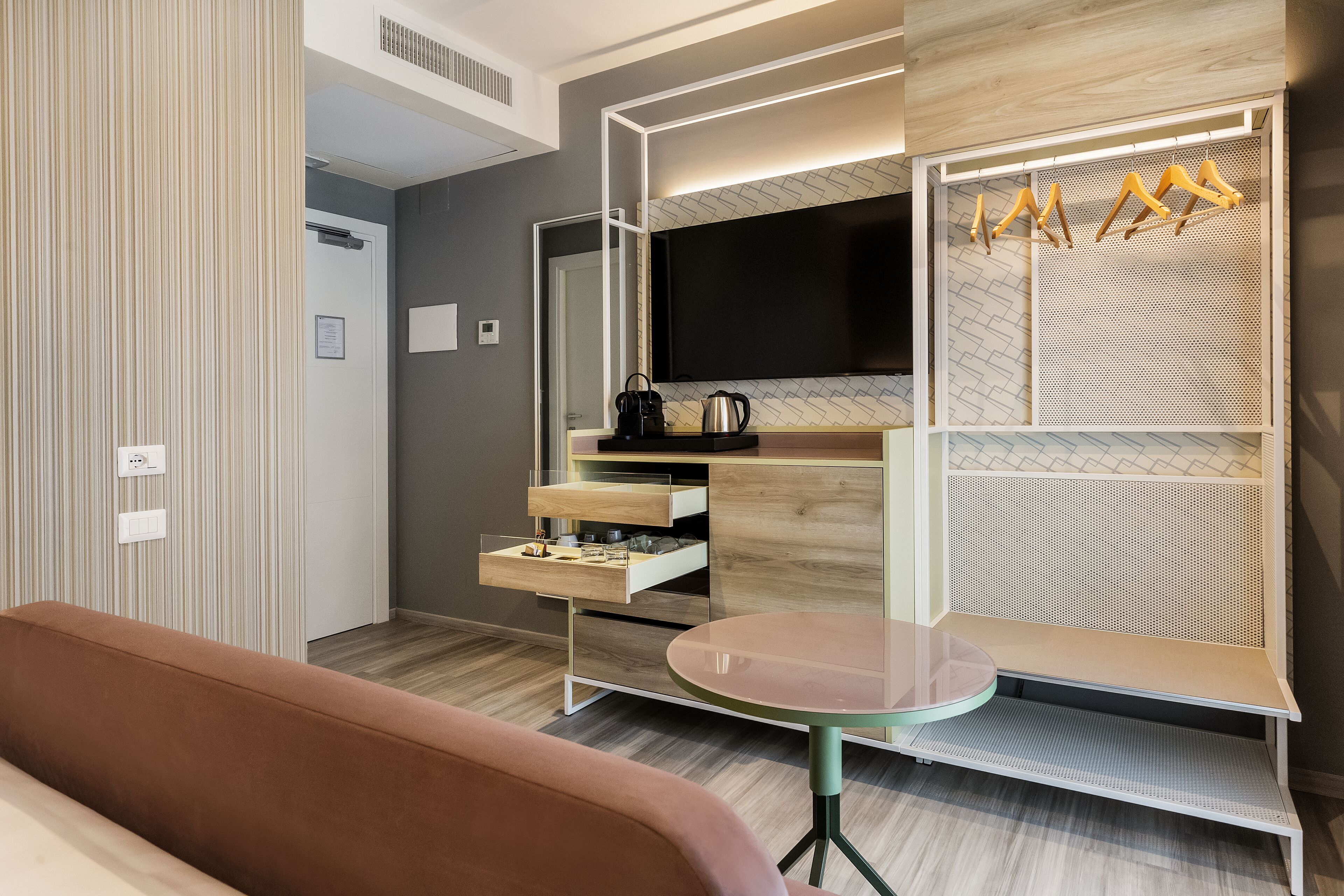 A modern hotel room with a wooden floor, a brown sofa, a round glass table, a TV mounted on a wooden panel, and a coat rack with hangers.