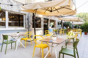 An outdoor dining area with yellow and green chairs, tables, and beige umbrellas.