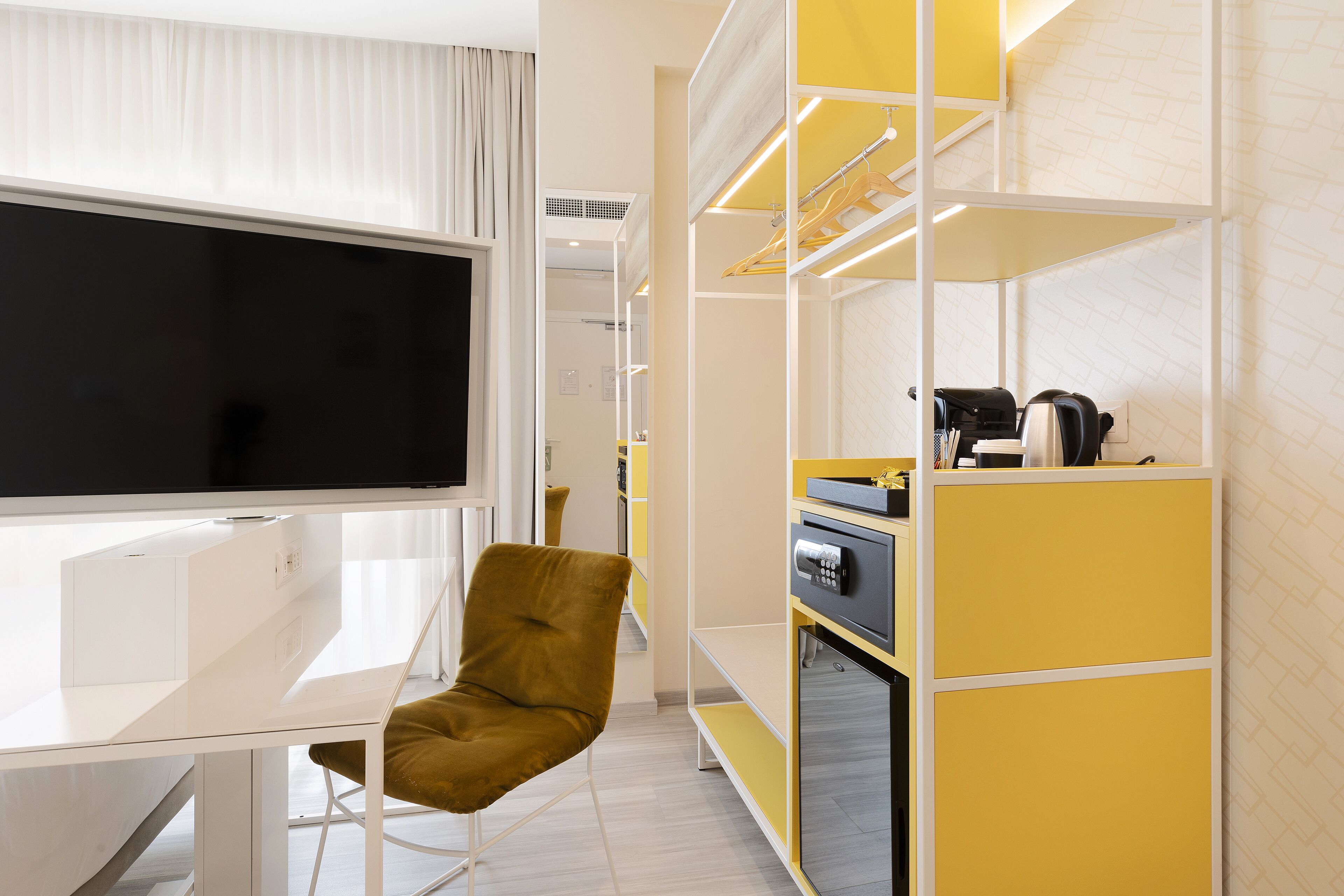 Una camera d'hotel moderna con una scrivania bianca, una sedia color senape, una grande TV a schermo piatto, un armadio con specchio e una macchina per il caffè.