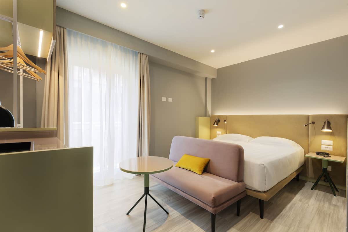 Una habitación de hotel con cama, un sofá con almohada amarilla, una mesita, un espejo y una mesita de noche con lámpara.