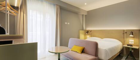 A hotel room with a bed, a sofa with a yellow pillow, a small table, a mirror, and a nightstand with a lamp.