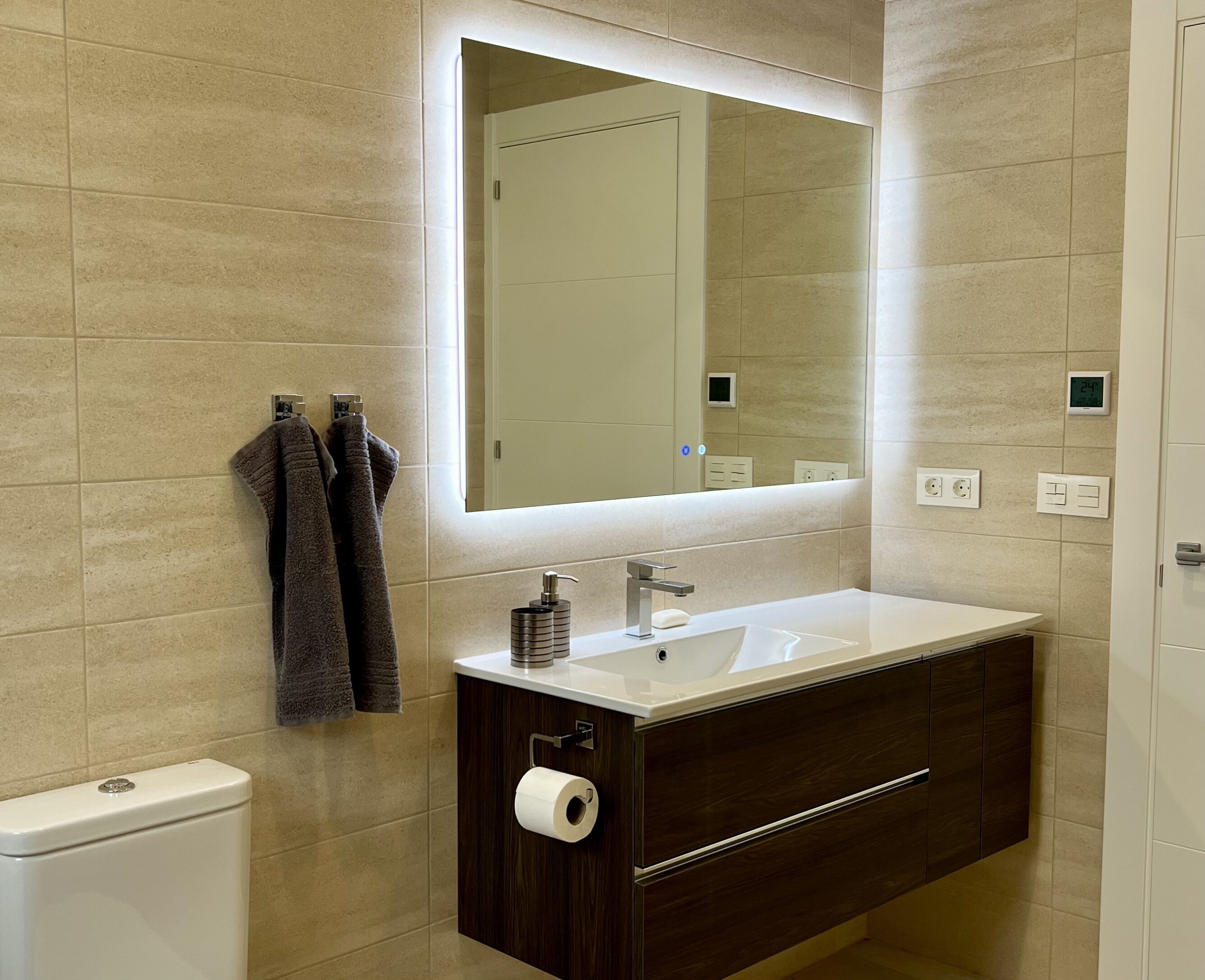 A modern bathroom with a large mirror, a white sink, a dark wooden cabinet, and a toilet.