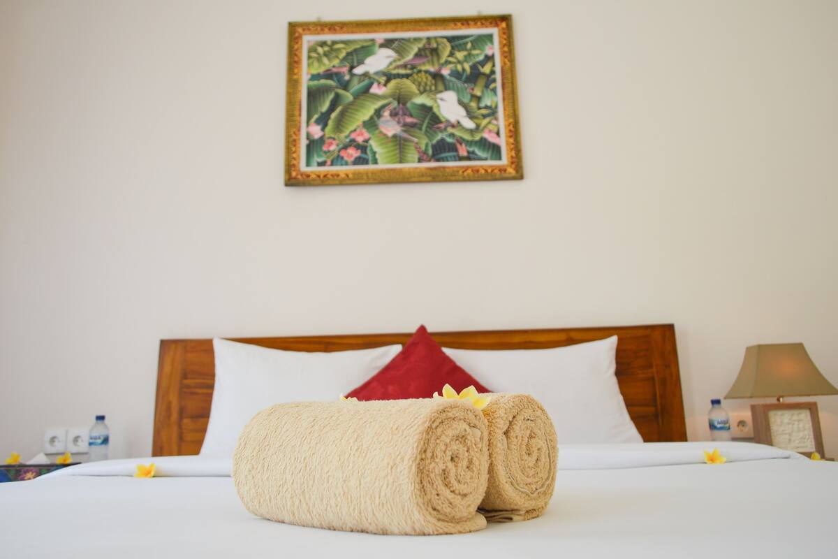 Una cama bien tendida con toallas enrolladas, un cabecero de madera, una almohada roja y un cuadro enmarcado en la pared.