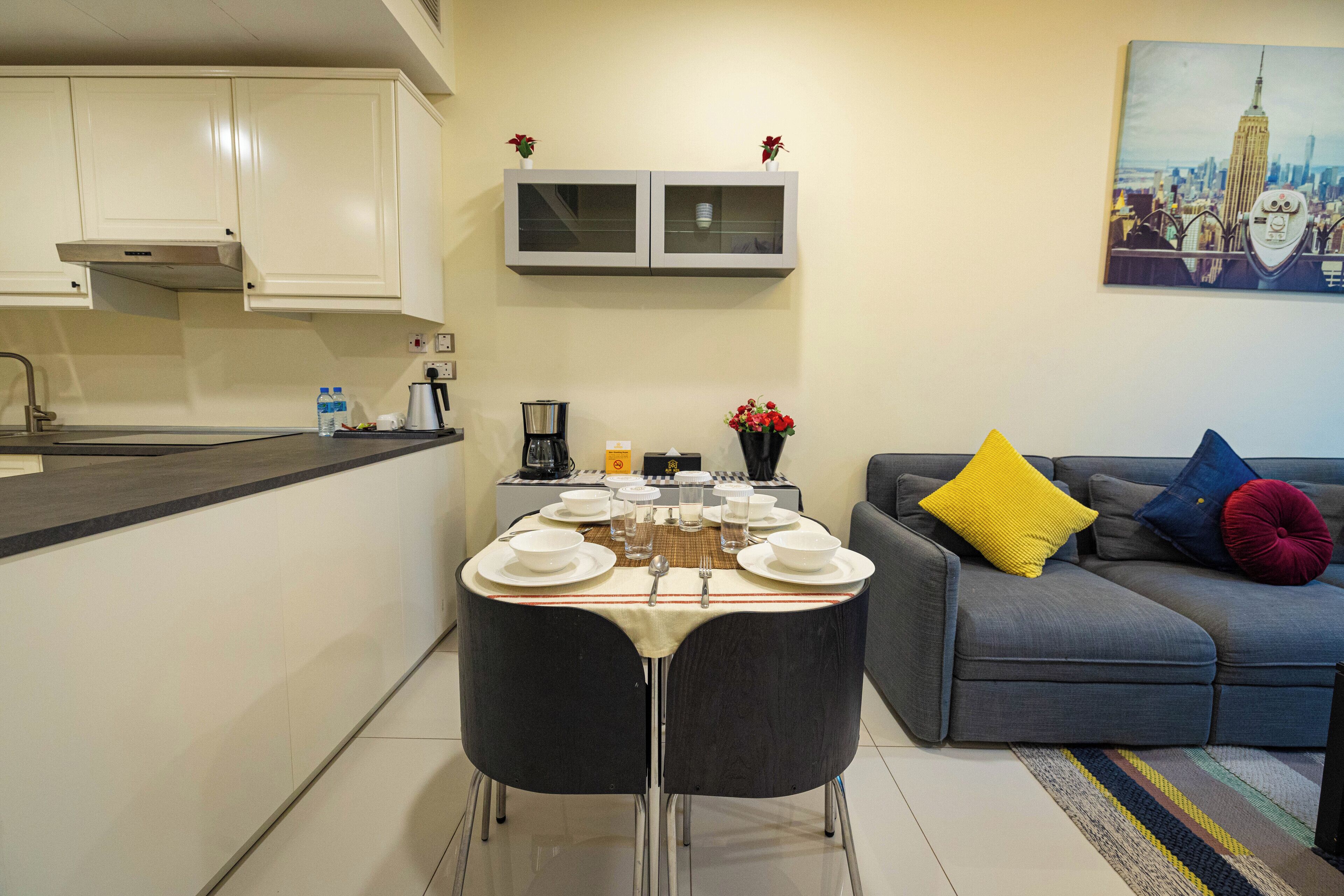 A modern kitchen with white cabinets, a dining table set for six with plates, glasses, and cutlery. A gray sofa with colorful cushions is in the living area, and a painting of a cityscape hangs on the wall.