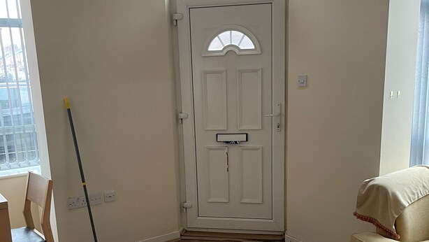 Ein Zimmer mit Holzboden, einer weißen Tür mit kleinem Fenster, einer Fußmatte, einem an die Wand gelehnten Besen und einem beigen Sofa.