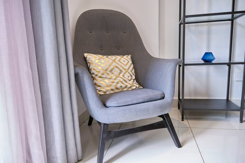 A grey armchair with a patterned cushion, positioned next to a metal shelving unit with a blue vase on top.