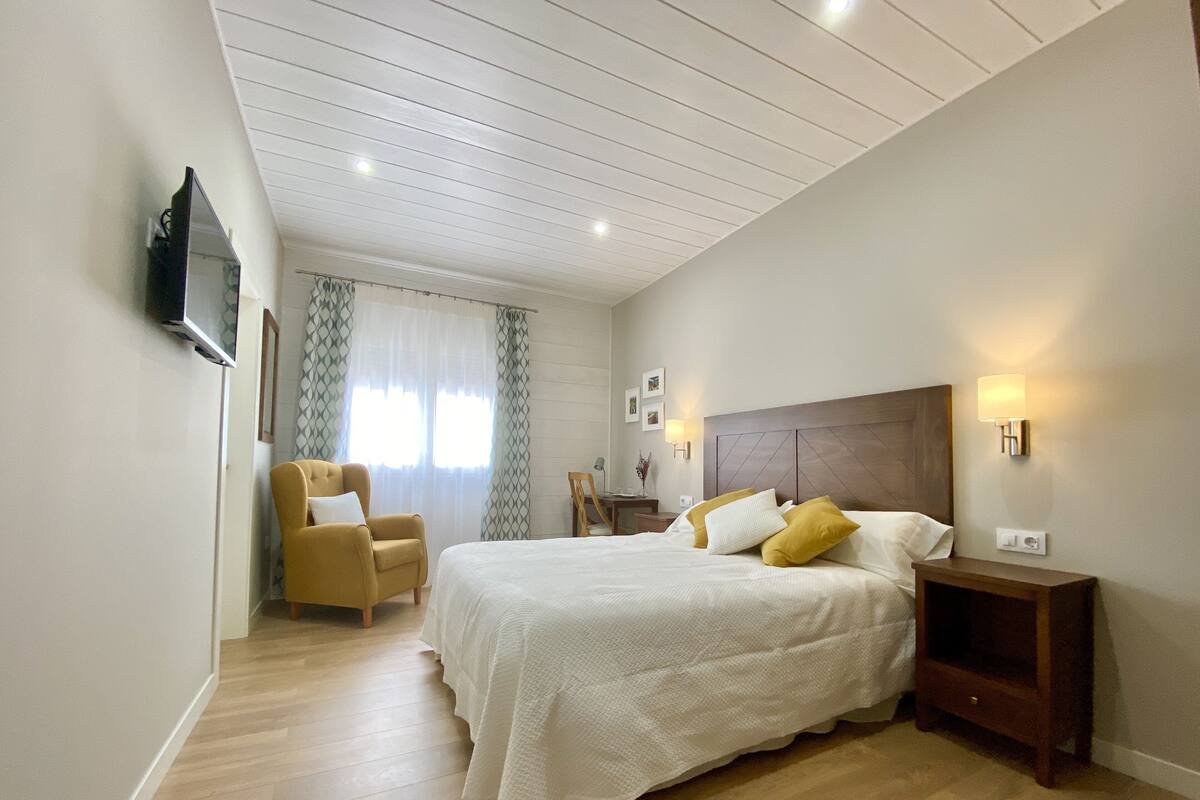 Un dormitorio con cama, una silla, una mesita de noche, televisor montado en la pared y una ventana con cortinas.