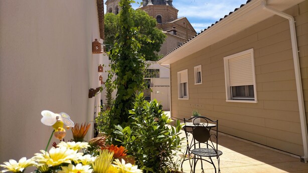 A narrow alleyway with a building featuring a bell tower and a small garden with flowers and a table and chair.