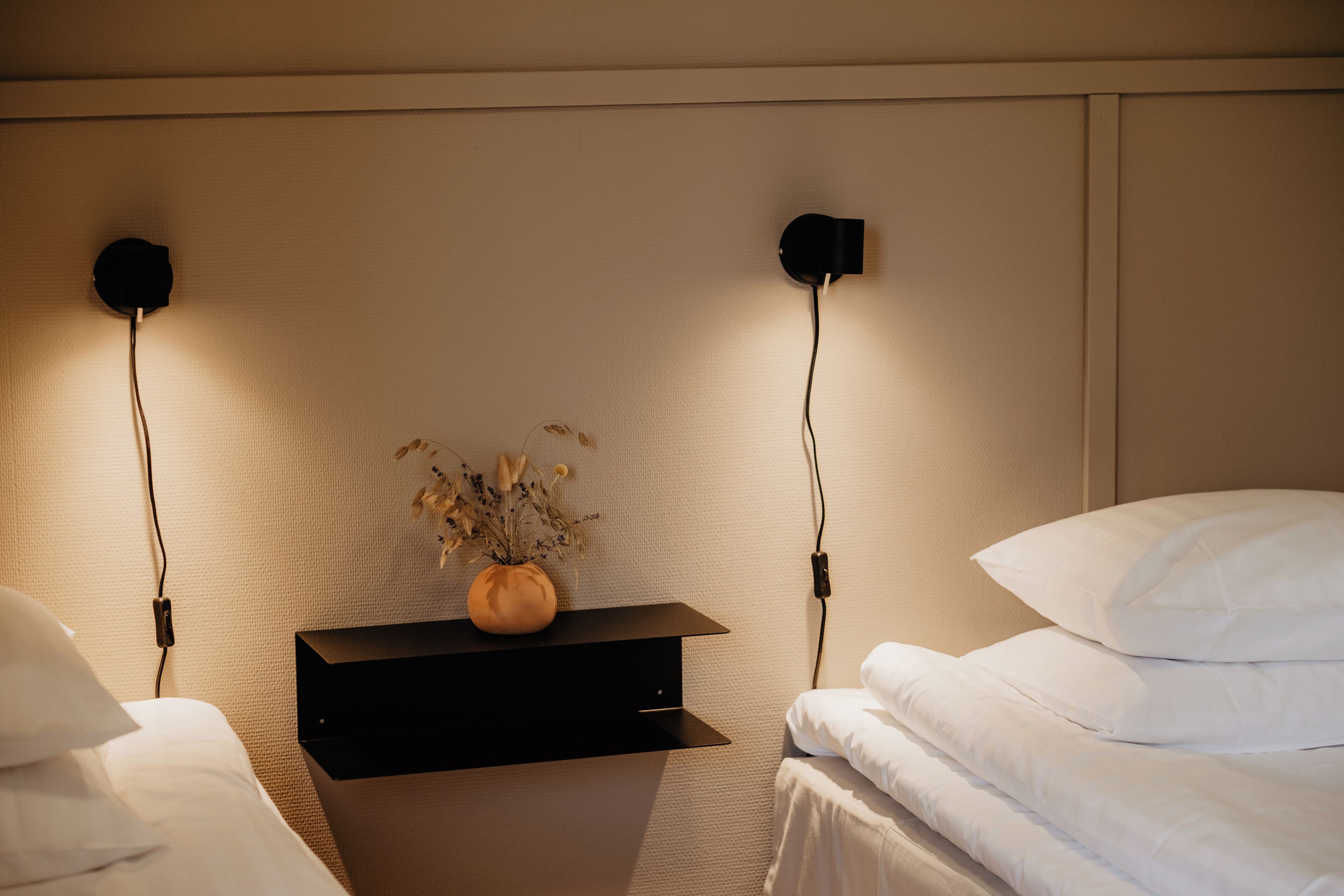 Una cama bien hecha con sábanas blancas, una mesita de noche con un jarrón de flores secas y dos luces de noche en la pared.